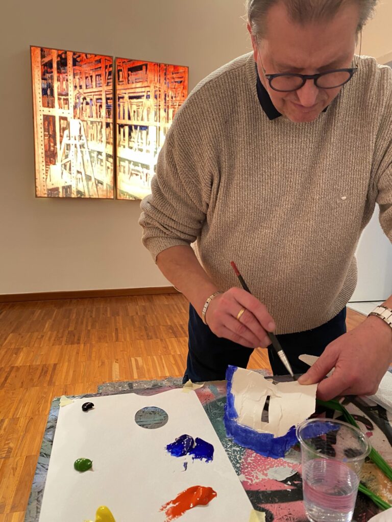Un uomo dipinge una maschera bianca su un tavolo da lavoro con colori acrilici, in una sala d’arte.