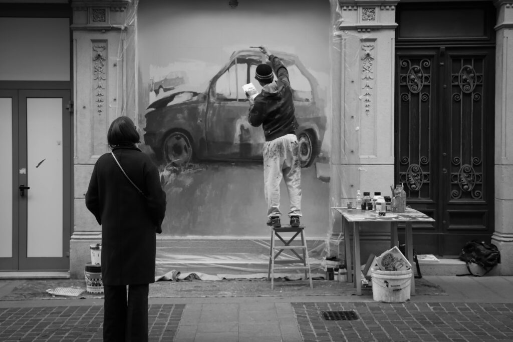 Fotografia in bianco e nero di un artista su uno sgabello che dipinge un murale di un'automobile su una parete esterna, osservato da una donna di spalle.
