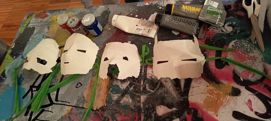 Quattro piccole maschere bianche fatte a mano, disposte in fila su un tavolo da lavoro macchiato di vernice. Ogni maschera ha occhi stilizzati ritagliati e fili verdi fissati come elementi decorativi. Attorno si vedono colori acrilici, tubetti di vernice e pennelli, suggerendo un’attività artistica in corso.