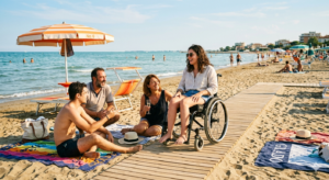 Un gruppo di amici si diverte sulla spiaggia accessibile di Grado, con una donna in sedia a rotelle che ride e un ombrellone a strisce arancioni e bianche.