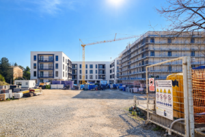 Cantiere edile sotto cielo sereno con complesso residenziale moderno: a sinistra edificio quasi completato, a destra struttura con impalcature, gru centrale e materiali da costruzione su piazzale ghiaioso illuminato dal sole.