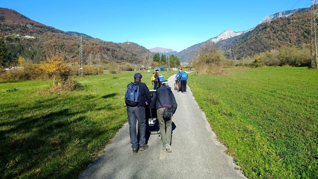 Un gruppo di persone percorre un sentiero asfaltato in una valle soleggiata, circondata da prati verdi e montagne, dirigendosi verso le vette in lontananza.