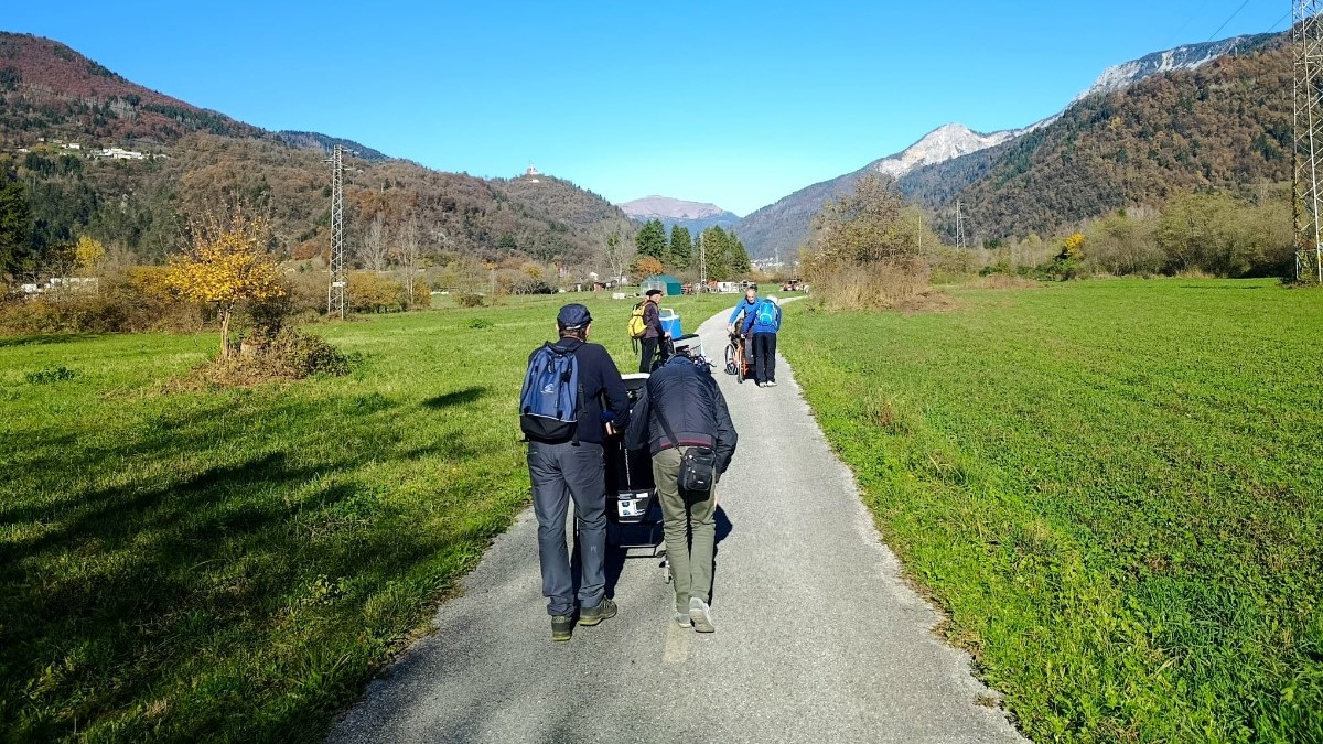 Un gruppo di persone percorre un sentiero asfaltato in una valle soleggiata, circondata da prati verdi e montagne, dirigendosi verso le vette in lontananza.