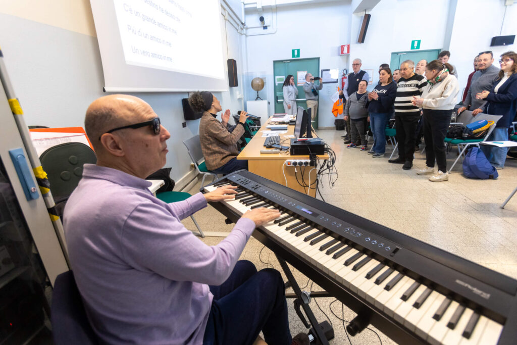 Mauro Costantini suona il piano elettrico mentre un gruppo di partecipanti, in piedi in fondo all'aula, accompagna la musica battendo le mani. Sullo schermo sono proiettati i testi di una canzone.