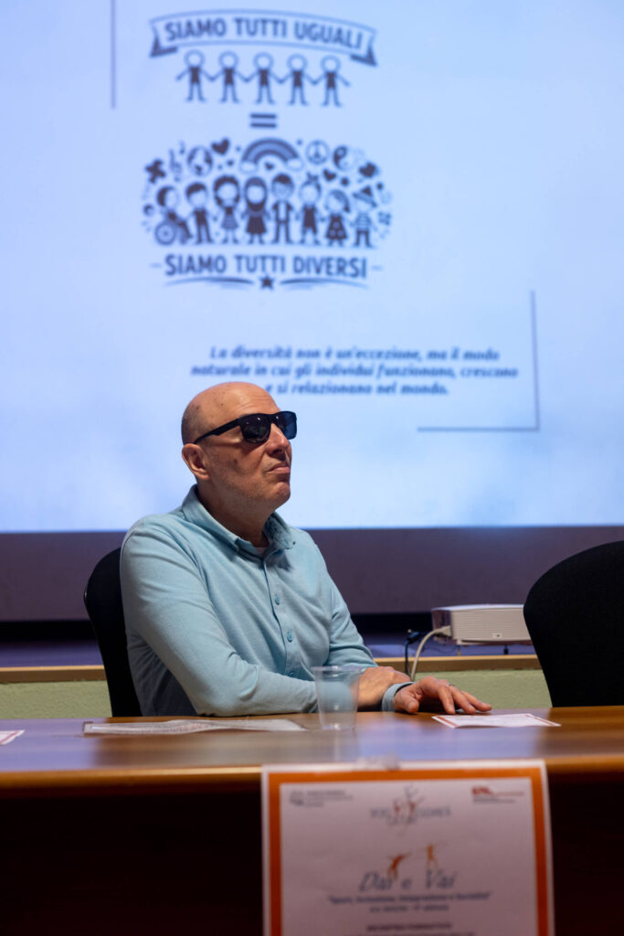 Mauro Costantini seduto al tavolo relatori davanti alla proiezione con la scritta "Siamo tutti uguali, siamo tutti diversi" e il volantino del progetto "Dai e vai".