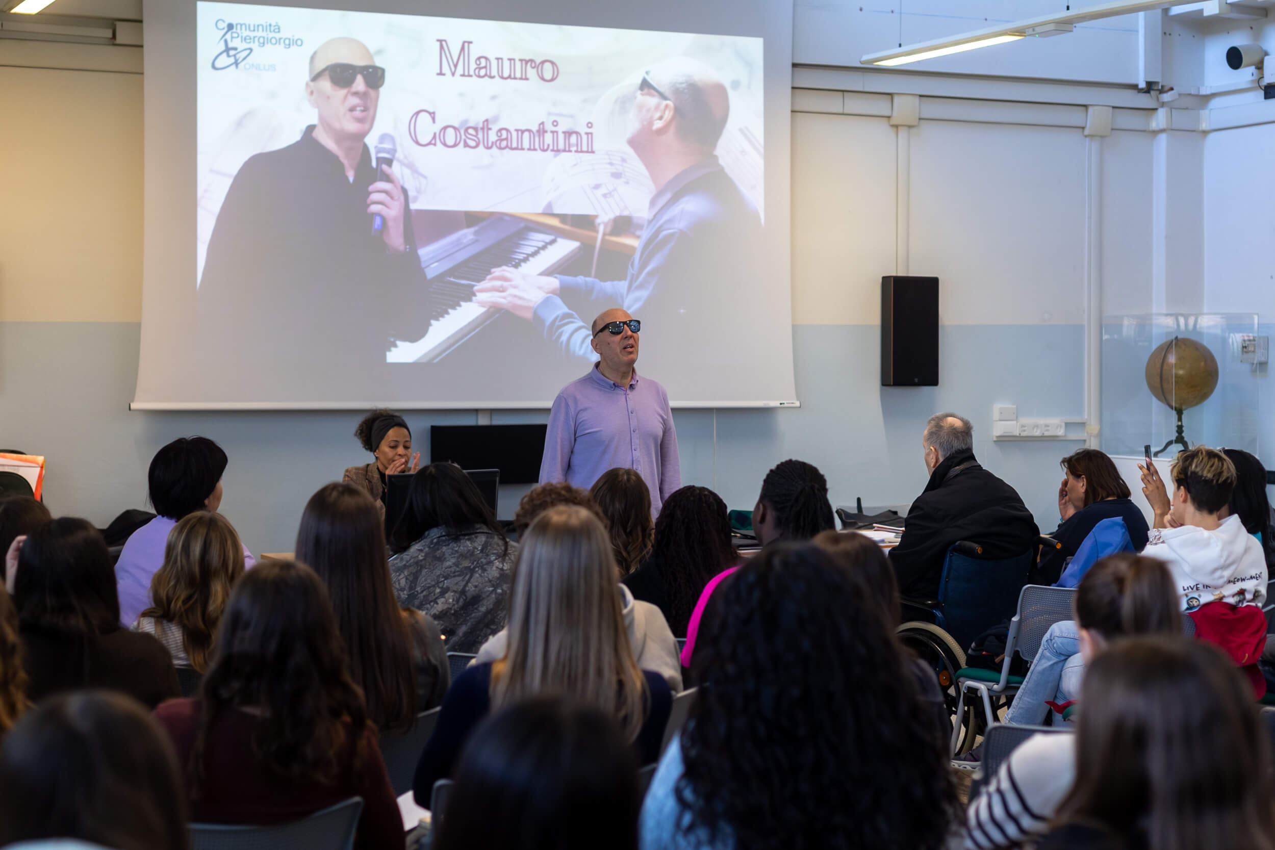 Mauro Costantini, in piedi davanti a una platea di studenti, durante un incontro del progetto "Dai e vai". Sullo sfondo, una presentazione proiettata con il logo della Comunità Piergiorgio ONLUS e immagini di Mauro mentre canta e suona il pianoforte.