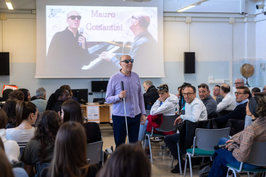 Mauro Costantini, con microfono e bastone bianco, parla in piedi tra il pubblico di studenti e adulti. Sullo schermo la presentazione della Comunità Piergiorgio.