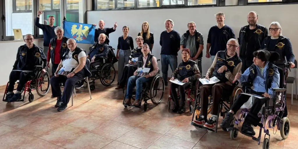Foto di gruppo al termine della lezione: allievi, maestri e operatori posano sorridenti in una sala luminosa. Due partecipanti espongono la bandiera del Friuli con l'aquila patriarcale.
