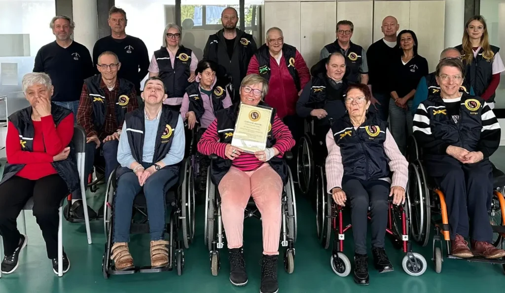 Foto di gruppo nel centro diurno: al centro un'allieva mostra con orgoglio l'attestato appena ricevuto. Intorno, il gruppo di allievi, maestri della Scuola Karate Udine e operatori della Comunità sorridenti.