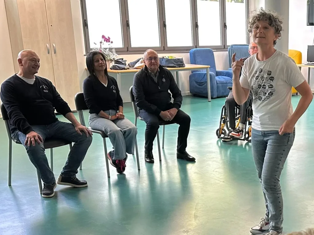 La cabarista Caterina Tomasulo, in arte Catine, in piedi sulla destra con una t-shirt bianca a fumetti, racconta uno dei suoi pezzi. A sinistra, seduti in ascolto, alcuni membri del pubblico sorridono divertiti.