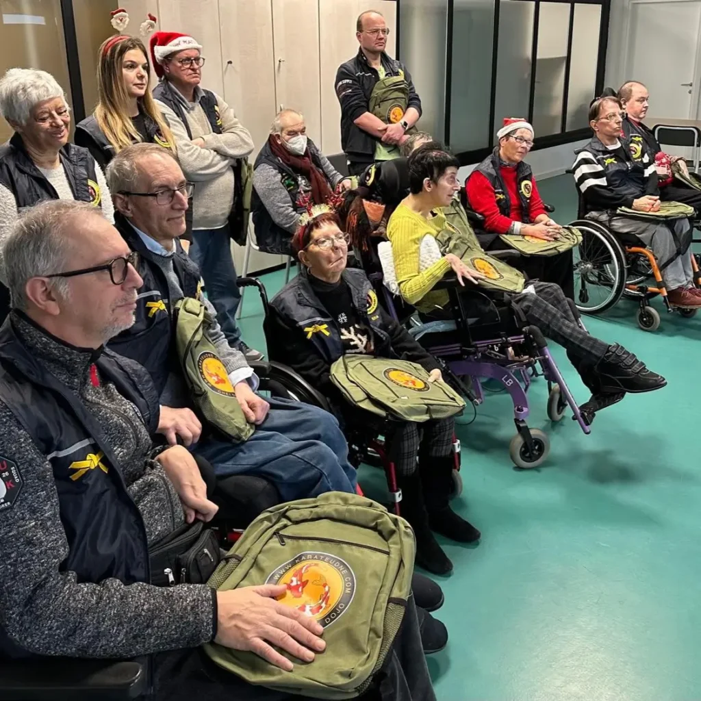 Gli allievi del corso, alcuni con cappellini natalizi, tengono in grembo degli zaini verdi con il logo di Karate Udine. Sono disposti in fila in un ambiente luminoso, in ascolto, come durante una consegna di piccoli regali.