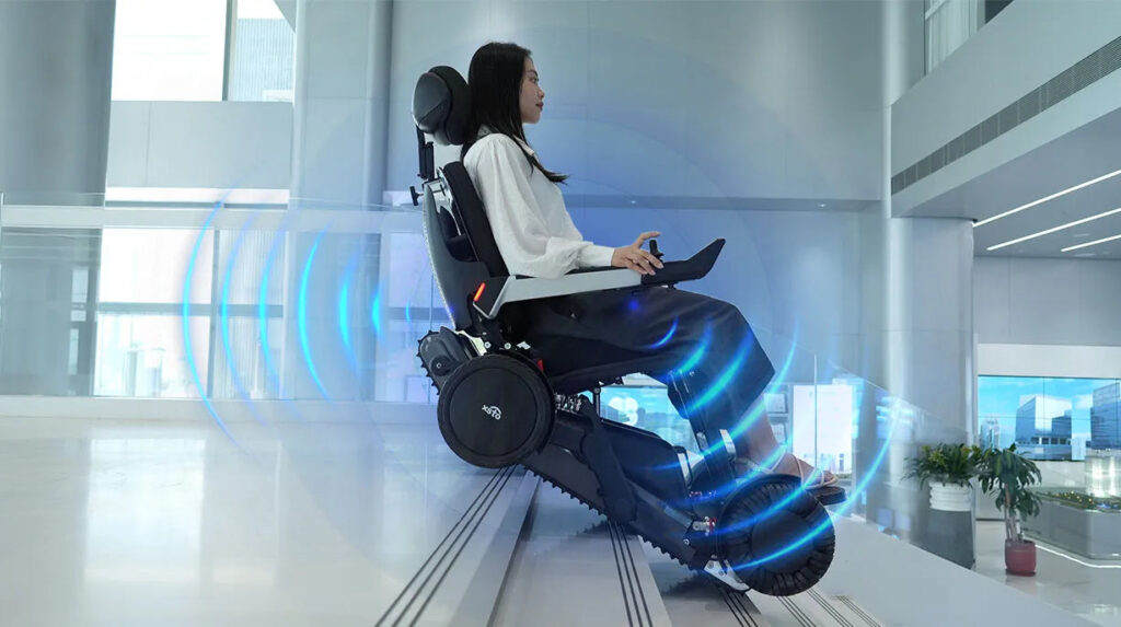 Persona seduta su una carrozzina elettrica X12 mentre scende una scala in un ambiente interno moderno, con effetti luminosi blu che suggeriscono tecnologia e controllo automatico.