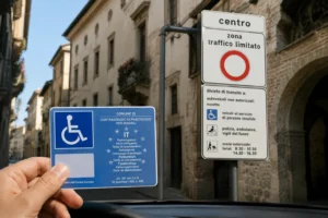 Una mano regge il contrassegno europeo di parcheggio per disabili davanti al cartello di ingresso a una Zona a Traffico Limitato.