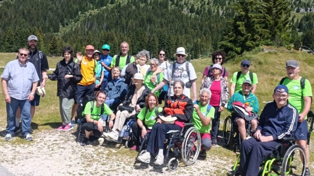Foto di gruppo dei partecipanti a Oltre le Vette, con persone in carrozzina e volontari in maglietta verde, sui prati del Montasio