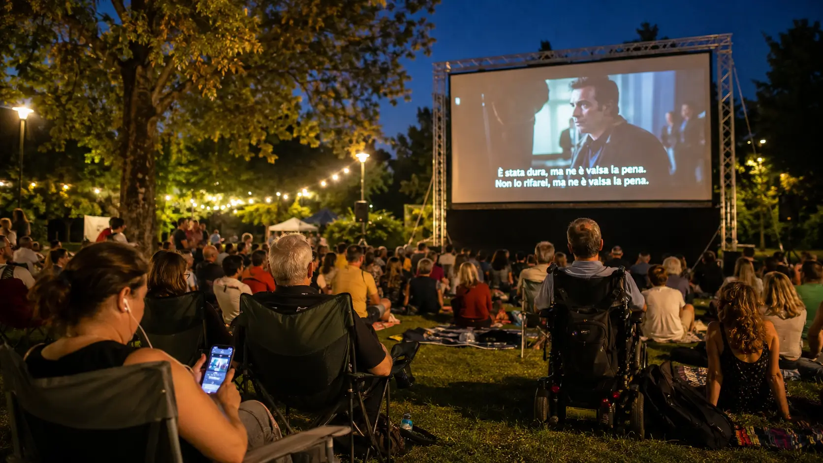 Pubblico seduto sul prato di un parco durante una proiezione serale all'aperto con sottotitoli sullo schermo; in primo piano una persona in carrozzina e una spettatrice con auricolari e smartphone.
