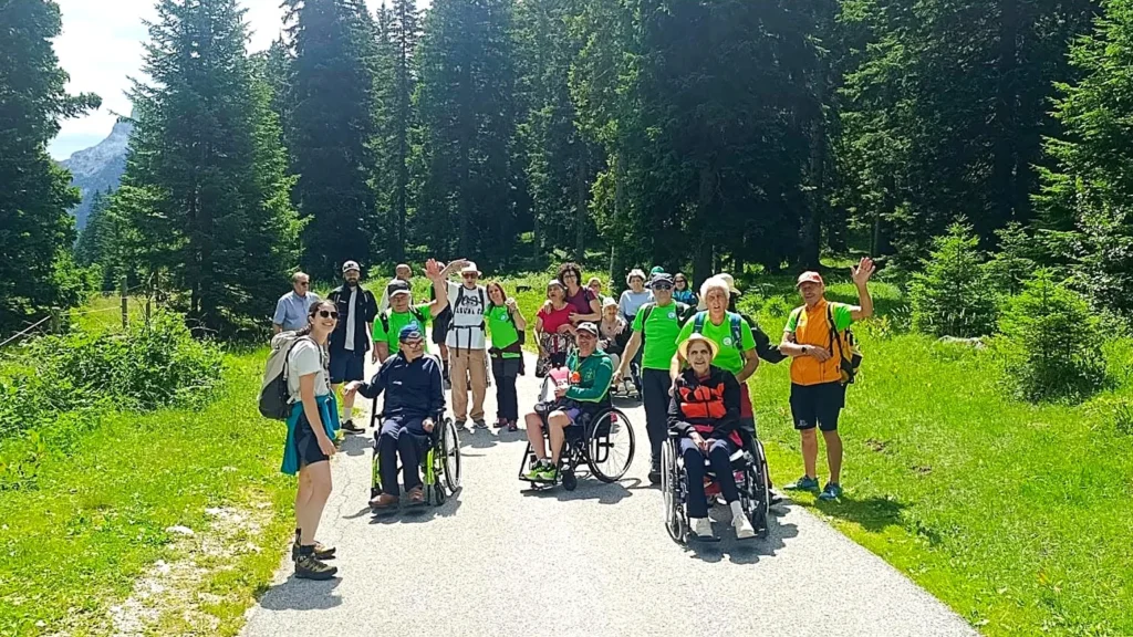 Gruppo di escursionisti con persone in carrozzina e volontari del CAI su un sentiero tra i boschi dell'Altopiano del Montasio