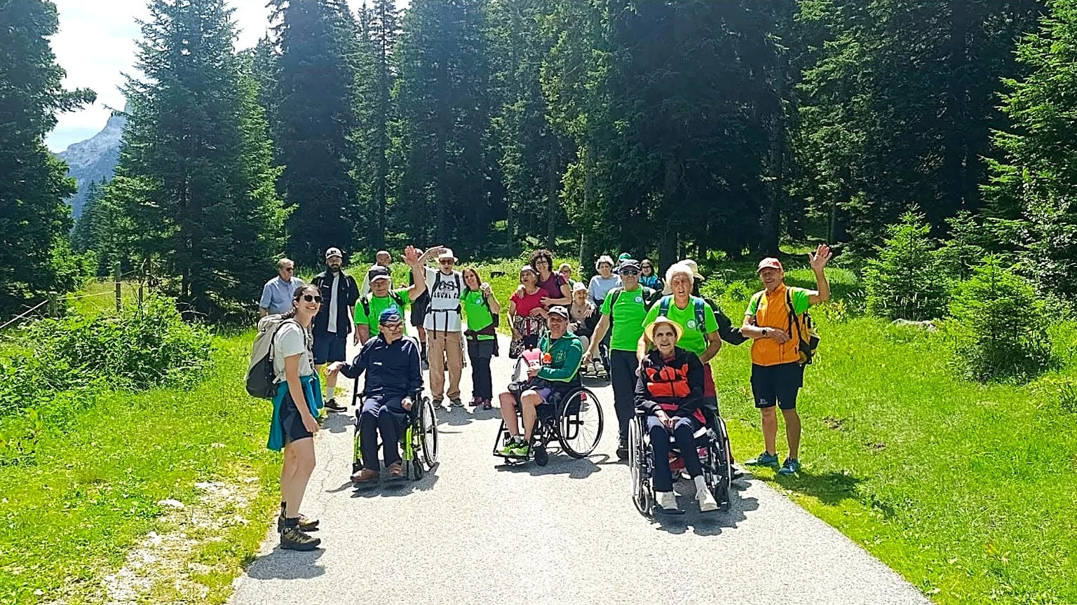 Gruppo di escursionisti con persone in carrozzina e volontari del CAI su un sentiero tra i boschi dell'Altopiano del Montasio
