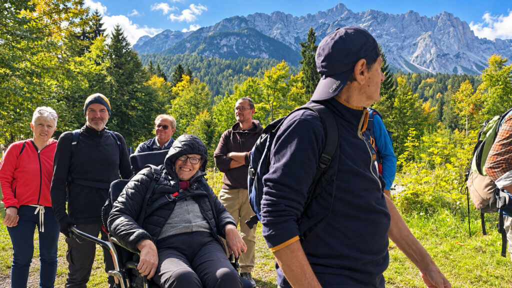 Gruppo di escursionisti in montagna accompagna una persona con disabilità su una joelette, lungo un sentiero immerso nel verde, con boschi e cime rocciose sullo sfondo.